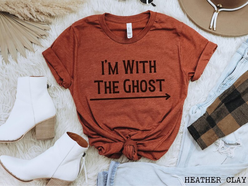 May include: A rust-colored t-shirt with the text "I'm with the ghost" and an arrow pointing to the right. The shirt is folded and displayed on a white surface.