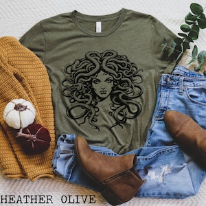 May include: A green t-shirt with a black and white graphic of Medusa, a mythical Greek figure with snakes for hair. The t-shirt is shown on a white surface with a brown sweater, blue jeans, and brown boots.