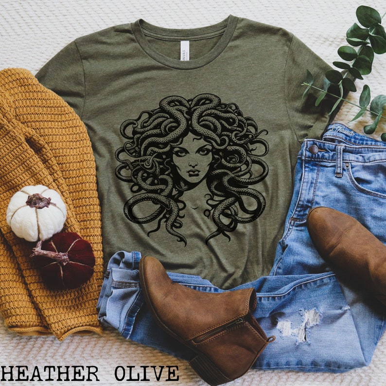 May include: A green t-shirt with a black and white graphic of Medusa, a mythical Greek figure with snakes for hair. The t-shirt is shown on a white surface with a brown sweater, blue jeans, and brown boots.