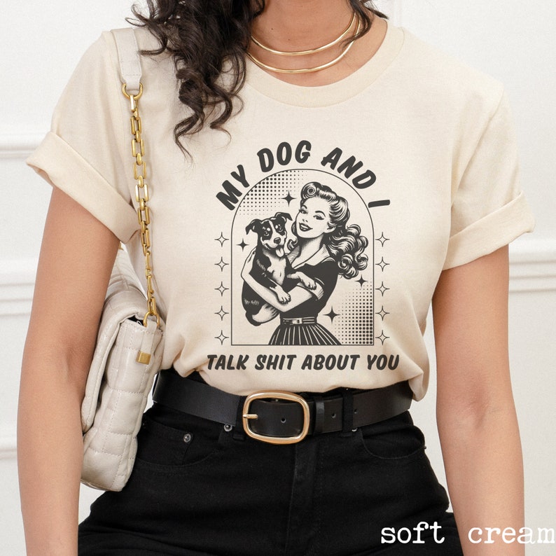 Op de afbeelding: Een cr&egrave;mekleurig T-shirt met een zwart-witte afbeelding van een vrouw die een hond vasthoudt. De afbeelding zegt "My Dog And I Talk Shit About You".