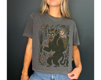 May include: Dark grey t-shirt featuring a Krampus illustration. The design shows a horned creature with a long tongue, carrying a child in a basket. The graphic is surrounded by stars and foliage. The shirt is a relaxed fit.