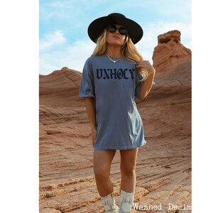 Op de afbeelding: Een vrouw draagt een blauw oversized T-shirt met het woord "UNHOLY" gedrukt in zwarte gotische letters. Ze draagt een zwarte hoed en een zonnebril en witte cowboylaarzen. De vrouw staat in een woestijnlandschap.