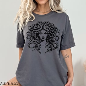 May include: A dark gray t-shirt with a black and white graphic print of Medusa, a mythical Greek figure with snakes for hair.