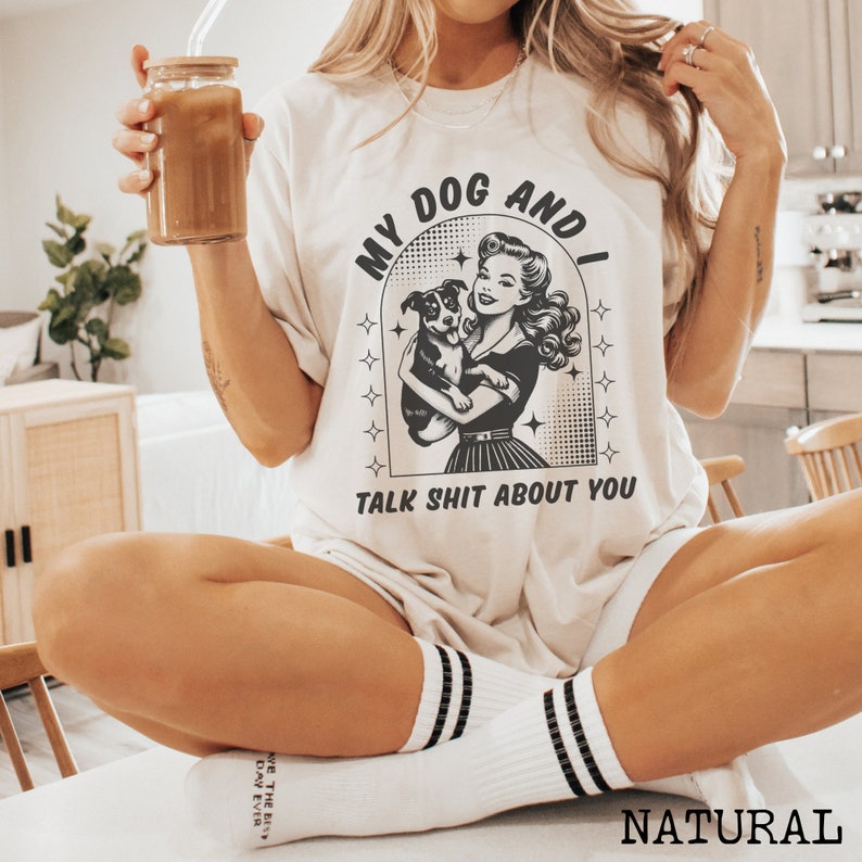 Op de afbeelding: Een wit T-shirt met een zwart-witte afbeelding van een vrouw die een hond vasthoudt. De afbeelding zegt "My Dog And I Talk Shit About You".