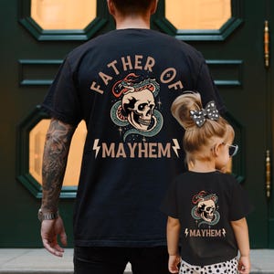 以下が含まれることがあります： 「Father of Mayhem」という文字と、蛇が巻き付いた頭蓋骨が描かれた黒いTシャツ。同じデザインの黒いTシャツを子供が着ています。