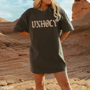 Op de afbeelding: Een oversized donkergrijs T-shirt met het woord "UNHOLY" gedrukt in een beige gotische letter.