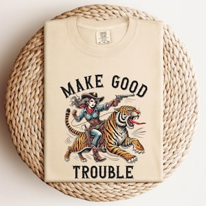以下が含まれることがあります： 虎に乗った女性が描かれたベージュのTシャツ。Tシャツには「Make Good Trouble」という文字がプリントされています。