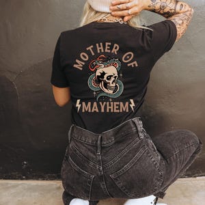 以下が含まれることがあります： 骸骨に蛇が巻き付いたグラフィックがプリントされた黒いTシャツ。骸骨の下に「Mother of Mayhem」という文字がプリントされています。