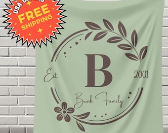 Könnte beinhalten: Ein hellgrüner Wandteppich mit einem braunen kreisförmigen Design mit dem Buchstaben "B", einem floralen Element und den Worten "Brink Family" und "Est. 2001". Der Wandteppich hängt vor einer weißen Backsteinmauer.