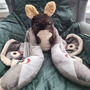 May include: A soft moth toy with grey wings and a brown body. The moth has large, black eyes and beige ears. The wings feature red and black details. The toy is placed on a green blanket.