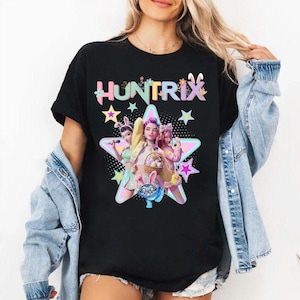 以下が含まれることがあります： 黒いTシャツ。カラフルな文字で「HUNTRIX」と書かれ、星の中にイースターバニーの格好をした3人の女性のグラフィックが描かれています。グラフィックには星、卵のバスケット、青いウサギが含まれています。