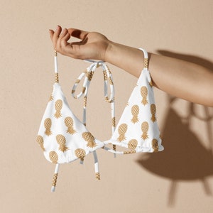 May include: White bikini top featuring a gold pineapple print. The triangle top has tie straps. The swimwear is held up by a hand against a neutral background.