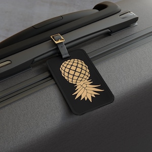May include: Black luggage tag with a gold pineapple design. The tag is attached to a black suitcase with a gold buckle and black strap. The pineapple design is a light gold color.