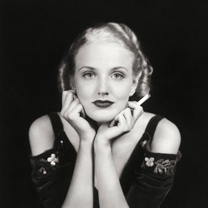May include: A black and white portrait of a woman with her chin resting on her hands. She is wearing a dark, off-the-shoulder dress with floral embellishments and has a cigarette in her hand.