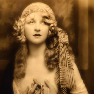 May include: A vintage sepia-toned photograph of a woman with long, wavy hair wearing a striped scarf and a pearl necklace. She is holding a small, ornate pendant in her hands.
