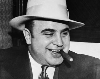 May include: Black and white portrait of a man wearing a light-colored fedora hat, a dark suit, and a patterned tie. He is smiling and holding a lit cigar in his mouth. The image is a medium shot, focusing on the man's face and upper body.