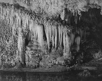 May include: Black and white photograph of a cave with stalactites and stalagmites. The cave is dark and the formations are illuminated by a light source.