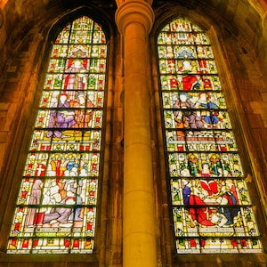May include: Two tall stained glass windows featuring colourful scenes of people in medieval attire. The windows are set within a stone archway with a thick pillar in the centre.