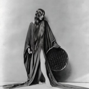 May include: A woman in a long, black robe stands with a woven basket in her hand. Her long, dark hair flows down her back. She is standing on a white background.