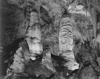 May include: Black and white photograph of a cave with tall, thin rock formations. The formations are made of stalactites and stalagmites.