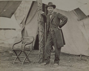 Pode incluir: Uma fotografia a preto e branco de um homem em uniforme militar de pé em frente a uma tenda. Ele está encostado em uma árvore e tem a mão na anca. Há uma cadeira em primeiro plano.