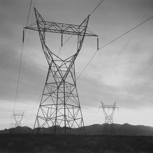 Op de afbeelding: Een zwart-witfoto van drie hoge, metalen elektriciteitstorens tegen een bewolkte lucht. De torens zijn geschetst tegen de bergen in de verte.