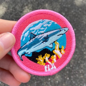May include: Pink embroidered patch featuring a blue shark swimming over coral reefs. The patch reads "Fiji" in yellow lettering.