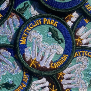 Könnte beinhalten: Nahaufnahme mehrerer gestickter Patches mit grünem Hintergrund, blauem Besatz und weißen Nähten. Die Patches zeigen ein Pilzdesign mit dem Text "Whitecliff Park" und "Kanada".
