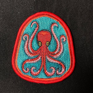 May include: Red embroidered octopus patch with pink tentacles on a teal background. The patch has a red border and is oval shaped.