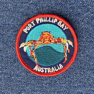 May include: Embroidered patch featuring a crab design with the text "PORT PHILLIP BAY AUSTRALIA". The patch has a red border, a blue background, and a crab in shades of orange and yellow. The patch is on a blue denim background.