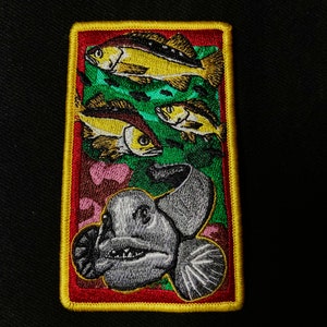 Könnte beinhalten: Ein gestickter Aufnäher mit einem grauen Fisch mit scharfen Zähnen und einem grünen Hintergrund mit gelben Fischen.