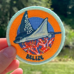 Peut inclure: Un écusson rond brodé représentant une raie manta bleue et blanche nageant au-dessus de coraux. L'écusson a un fond orange avec un cercle bleu au centre. Le texte "Belize" est écrit en blanc en bas de l'écusson.