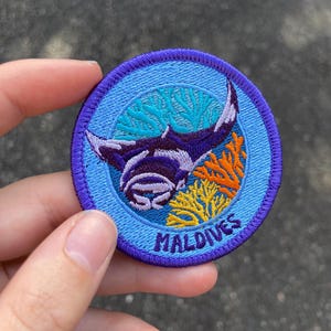 May include: A blue and purple embroidered patch featuring a manta ray swimming over coral reefs. The patch reads "MALDIVES" in yellow lettering.