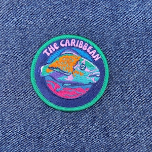 Puede incluir: Un parche redondo bordado con el texto "The Caribbean". El parche presenta un diseño de pez colorido en tonos naranja, morado y rosa, sobre un fondo azul con un borde verde. El parche está sobre un fondo de mezclilla azul.