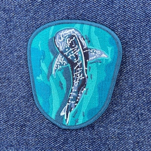 以下が含まれることがあります： ターコイズブルーの水の中を泳ぐジンベエザメの刺繍パッチ。青と黒のディテールが施されています。