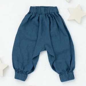 Puede incluir: Un pantalón de bebé de lino azul con cintura elástica. El pantalón está colocado sobre una superficie blanca con formas de estrellas y medias lunas de madera.