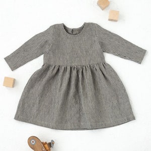 Op de afbeelding: Een grijs en wit gestreepte jurk met lange mouwen. De jurk heeft een ronde halslijn en een gerimpelde rok. Houten speelgoedblokken en een houten vliegtuig liggen verspreid rond de jurk op een wit oppervlak.