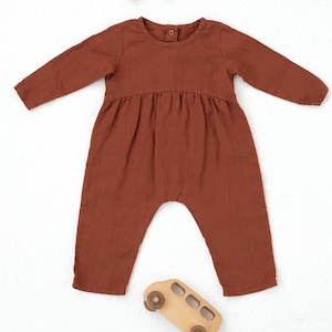 Könnte beinhalten: Ein rostfarbener Baby-Strampler mit langen Ärmeln und geraffter Taille. Das Outfit ist aus weichem Stoff gefertigt und hat einen Knopfverschluss am Rücken. Ein Holzspielzeugbus und kleine Holzklötze sind ebenfalls im Bild.