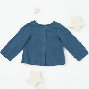 Peut inclure: Une chemise bleue à boutons et à manches longues, probablement pour un enfant. La chemise est en tissu doux et a un col rond. Des formes décoratives d'étoiles et de cylindres entourent la chemise.