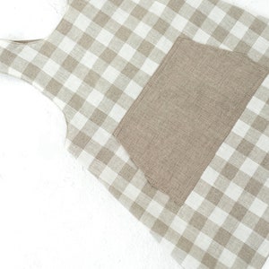 May include: A beige and white gingham apron with a large patch pocket. The apron has a sleeveless design and is made of a linen fabric.