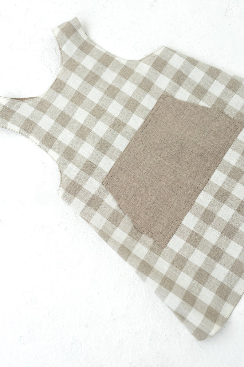 May include: A beige and white gingham apron with a large patch pocket. The apron has a sleeveless design and is made of a linen fabric.