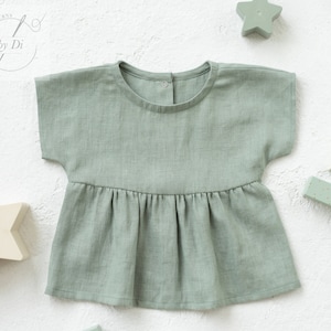 Pode incluir: Uma blusa de bebê de linho verde-sálvia com mangas curtas e saia franzida. A blusa tem um decote redondo e um fecho de botão nas costas.