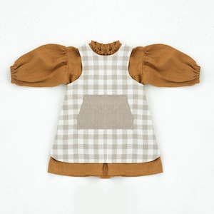 May include: A brown and white gingham dress with a brown linen top and long sleeves. The dress has a large pocket on the front.