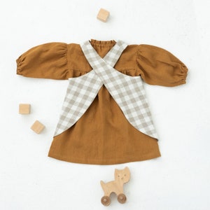 May include: A brown linen dress with a white and brown gingham overlay. The dress has a criss-cross back and long sleeves. There are wooden blocks and a wooden toy cat on the white background.