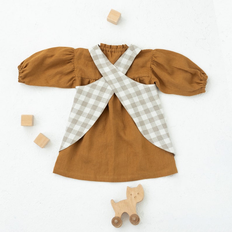 May include: A brown linen dress with a white and brown gingham overlay. The dress has a criss-cross back and long sleeves. There are wooden blocks and a wooden toy cat on the white background.