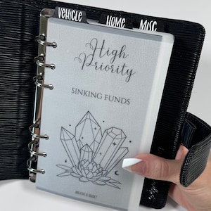 Puede incluir: Una carpeta negra con una página blanca que dice "High Priority Sinking Funds". La página tiene un dibujo de cristales y una flor. La carpeta tiene pestañas que dicen "Vehicle", "Home", y "Misc."