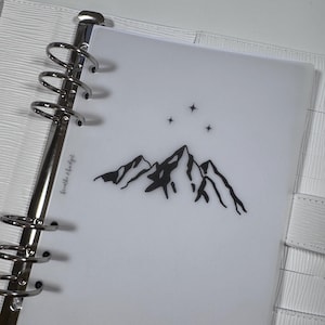 Puede incluir: Una agenda blanca con un inserto de plástico transparente que presenta una silueta negra de montañas y estrellas. El texto "breathe & budget" está impreso en la agenda.