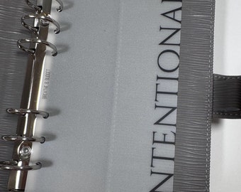 May include: A gray leather bound planner with a clear plastic insert that says "INTENTIONAL" in black text. The planner has a ring binder system with six rings.