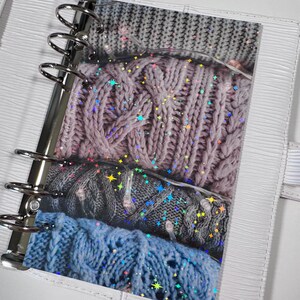 Puede incluir: Una carpeta blanca con un bolsillo de plástico transparente que contiene una pila de muestras de tejido de punto. El tejido es en tonos de gris, rosa y azul. El bolsillo de plástico tiene un patrón holográfico de estrellas.