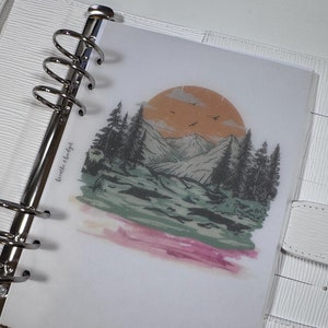 May include: A white planner with a clear plastic sheet insert featuring a watercolor illustration of a mountain range with a sunset behind it. The text "breathe & budget" is written on the sheet.